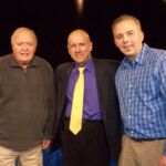 On the set of 'Beyond the Game'. Left to Right - Phil Lilley, John Vorperian (Host), Andrew Lilley (Filmmaker) Three men standing next to each other in front of a blue wall.